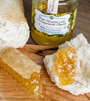 Jar of raw acacia honey in the background.  White bread and honeycomb chunk on wooden board in the foreground.