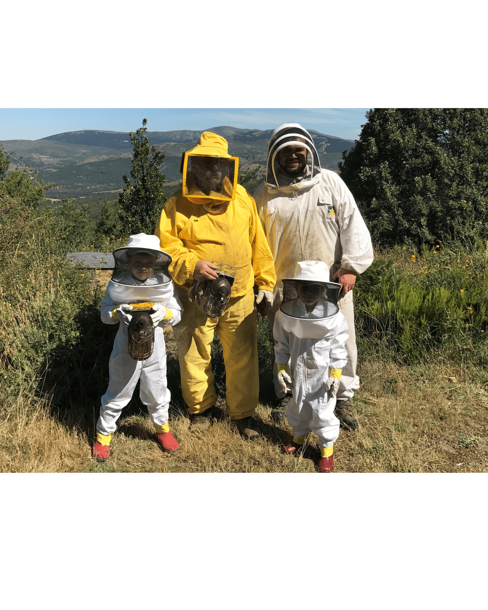 Family of four beekeepers wearing beekeeper suits comprising two adults and two children.